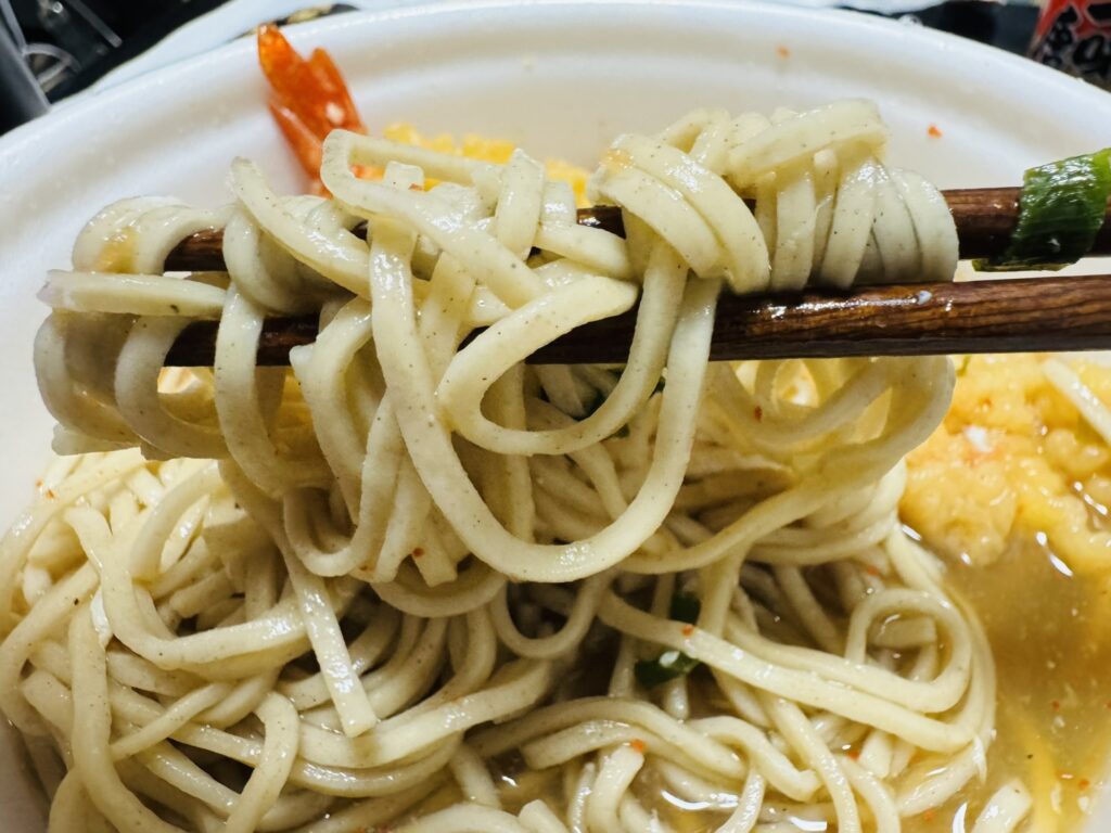 箸で麺を持ち上げた箸上げ写真。つゆと具材の一部が背景に見えている。