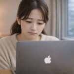 冬の室内で、MacBookを見つめながら残念そうな表情を浮かべる女性。