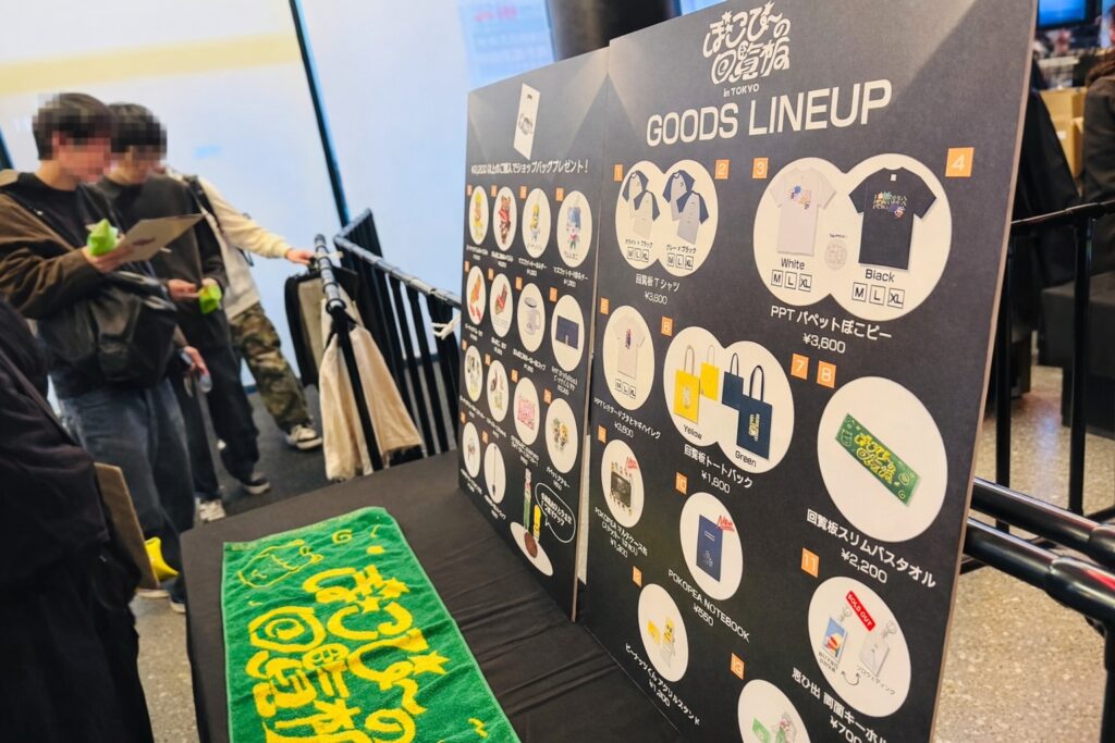 物販コーナーの『GOODS LINEUP』パネルとグッズ展示（タオルなど）